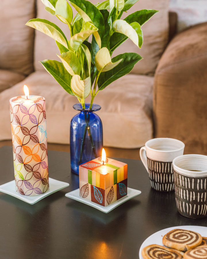 Lifestyle photo of Magic Garden 3x8 pillar and Magic Garden 3x3x3  cube candle, both lit and sitting on white candle coasters on a table in a casual living room setting.  Lovely pokes of color and light. 