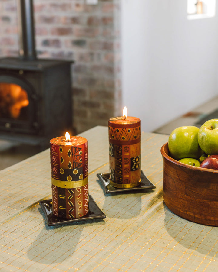 Lifestyle photo of 2 Safari Gold 3x6 pillars, lit and both on black candle coasters. On a table with the fire burning in the background.  Lovely tones of brown and rust with gold detail.