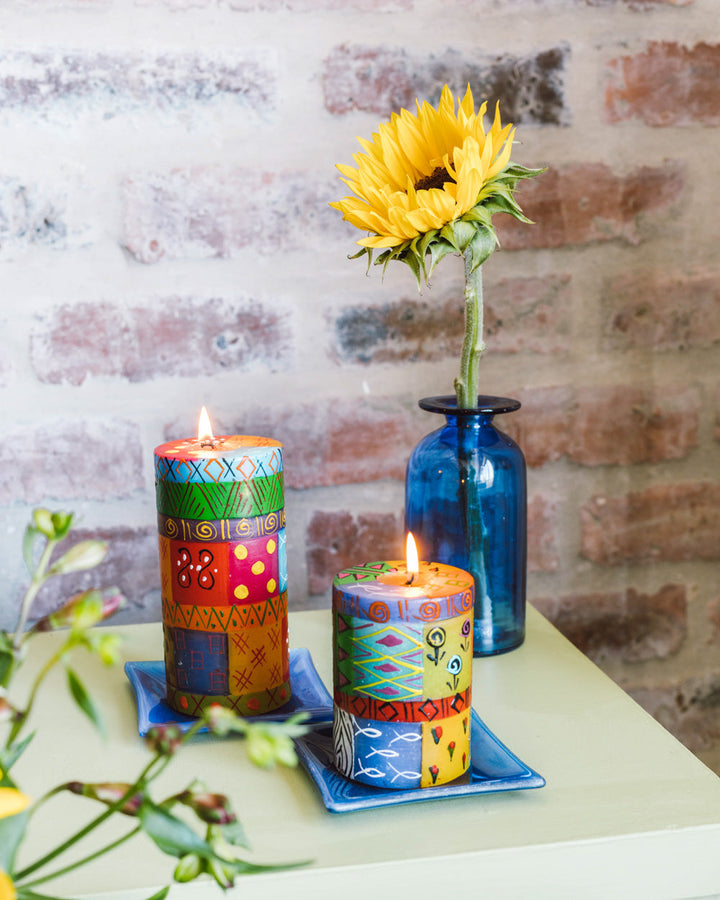 Lifestyle photo of Multi Color Ethnic 3x6 pillar and 3x4 pillar.  Both on deep blue candle coasters and beautifully burning!  Blue vase with a sunflower on table alongside.