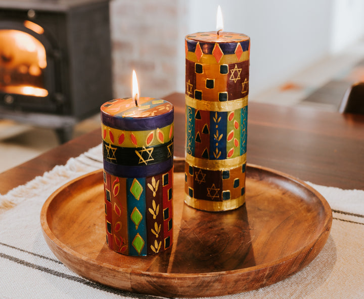 Lifestyle photo of the Judaica 3x6 pillar alongside the 3x8 pillar, both burning brightly on a wooden plate as the table center piece.  Fire burning in the background.  Beautiful!