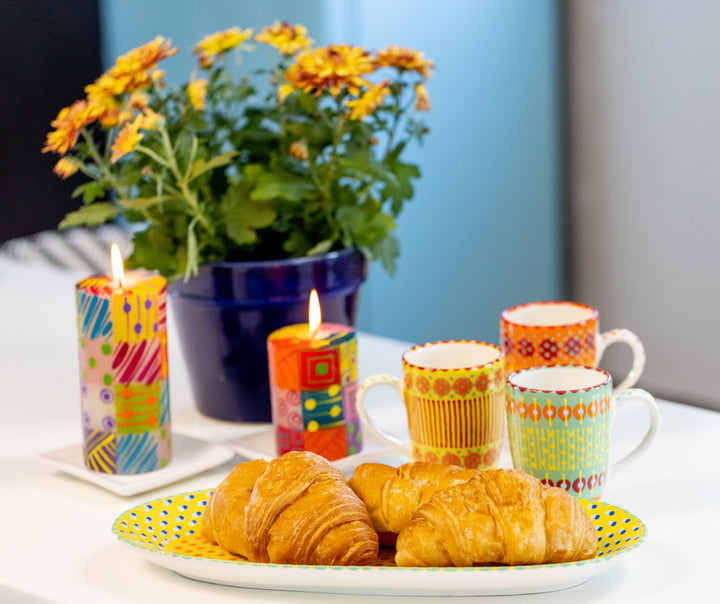 Lifestyle photo of three mugs; yellow, japer and orange alongside a yellow platter with croissants.  Burning beside this are 2 carousel pillars with flowers in the background  Colorful table setting!, 