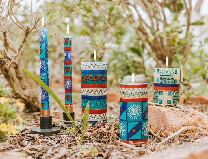 Lifestyle photo of African Sky tapers and pillars outside and brightly burning! Turquoise, dark blue, cream and dark tan in dots, sunbursts, and geometric designs.